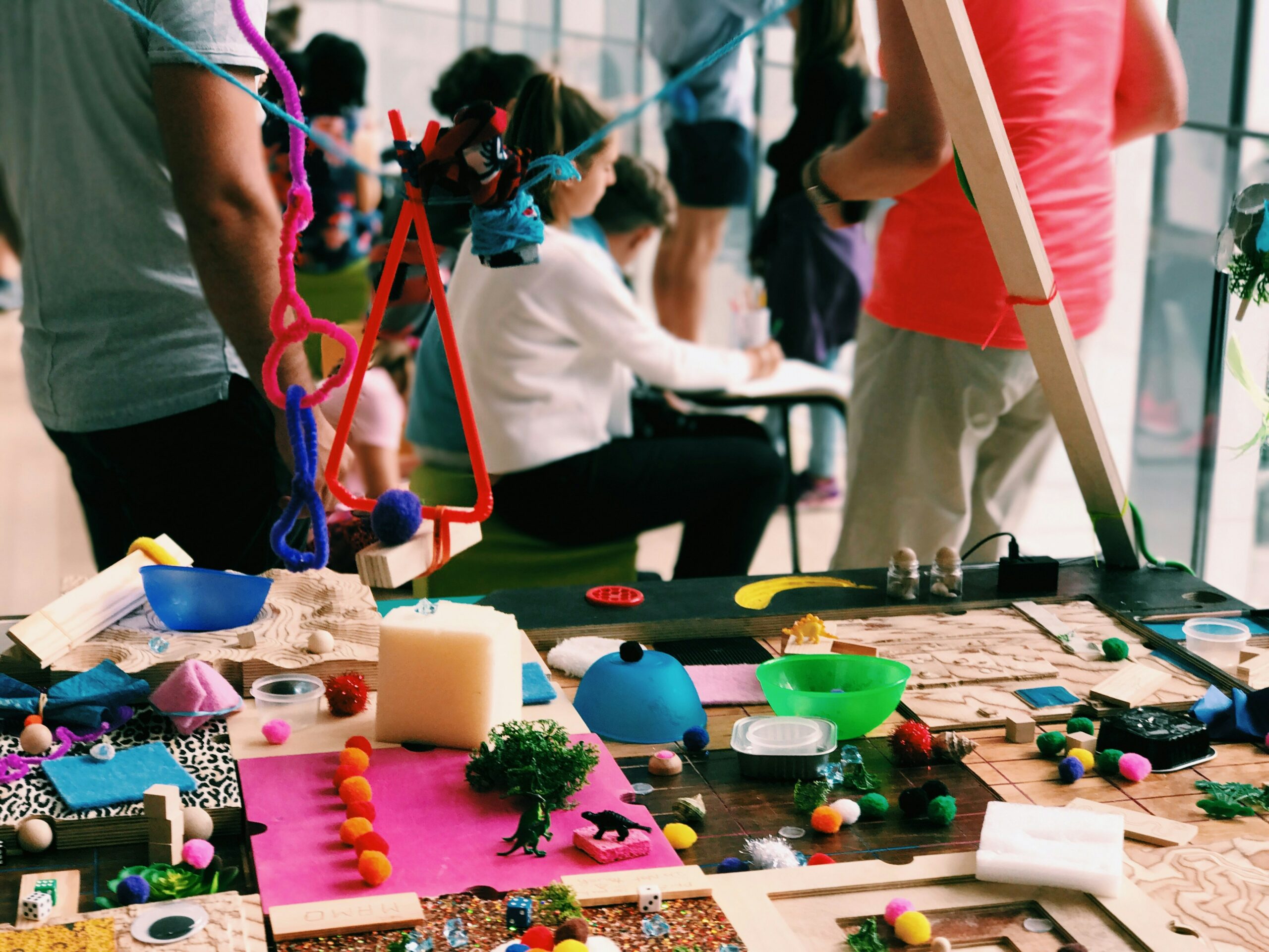 A colorful and vibrant art workshop setup featuring various crafting materials on a table, including pom-poms, pipe cleaners, fabric swatches, foam blocks, wooden pieces, and small decorative items.