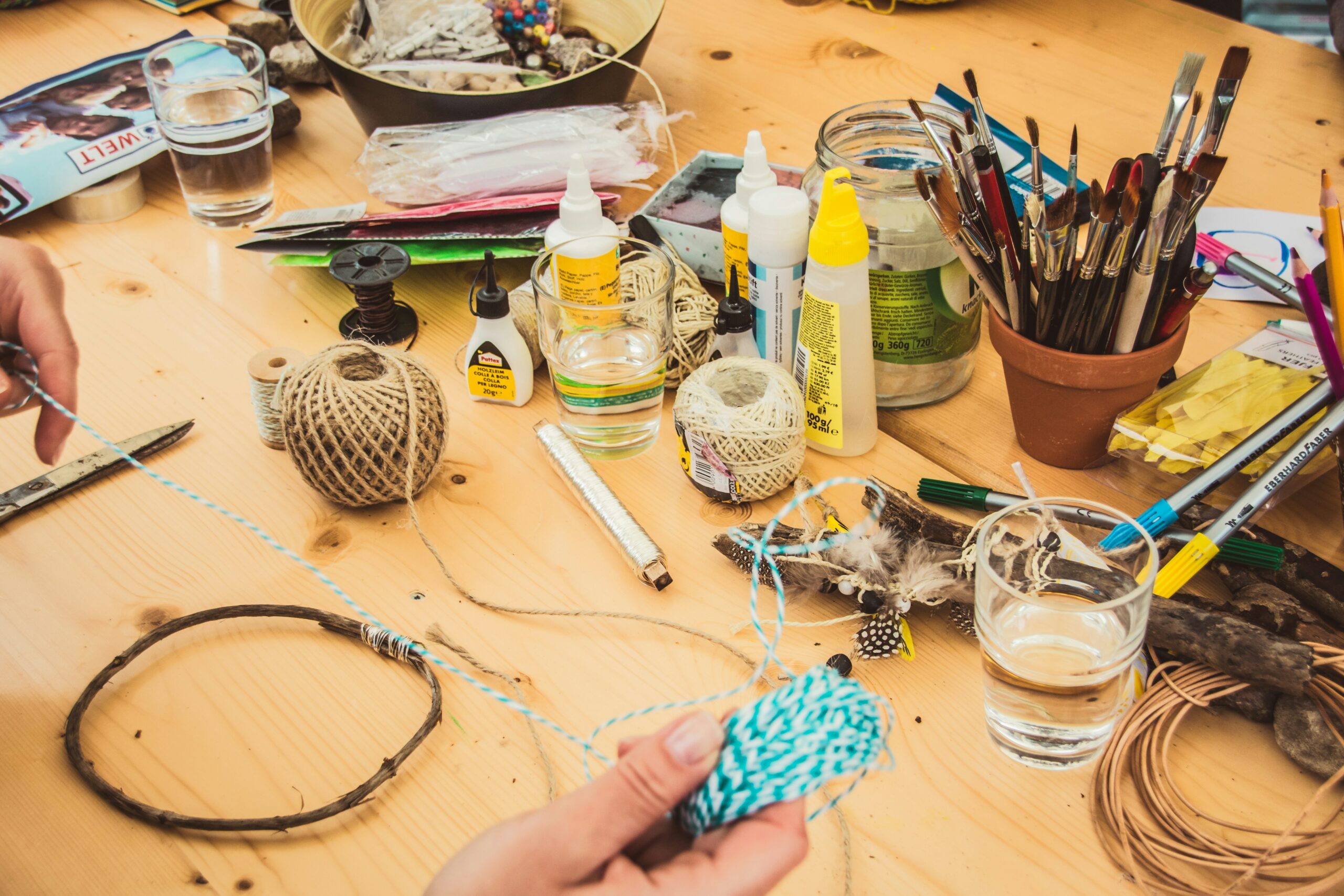 A crafting workspace with materials like twine, scissors, glue, feathers, and paintbrushes. A hand is seen handling blue string while assembling a craft project.
