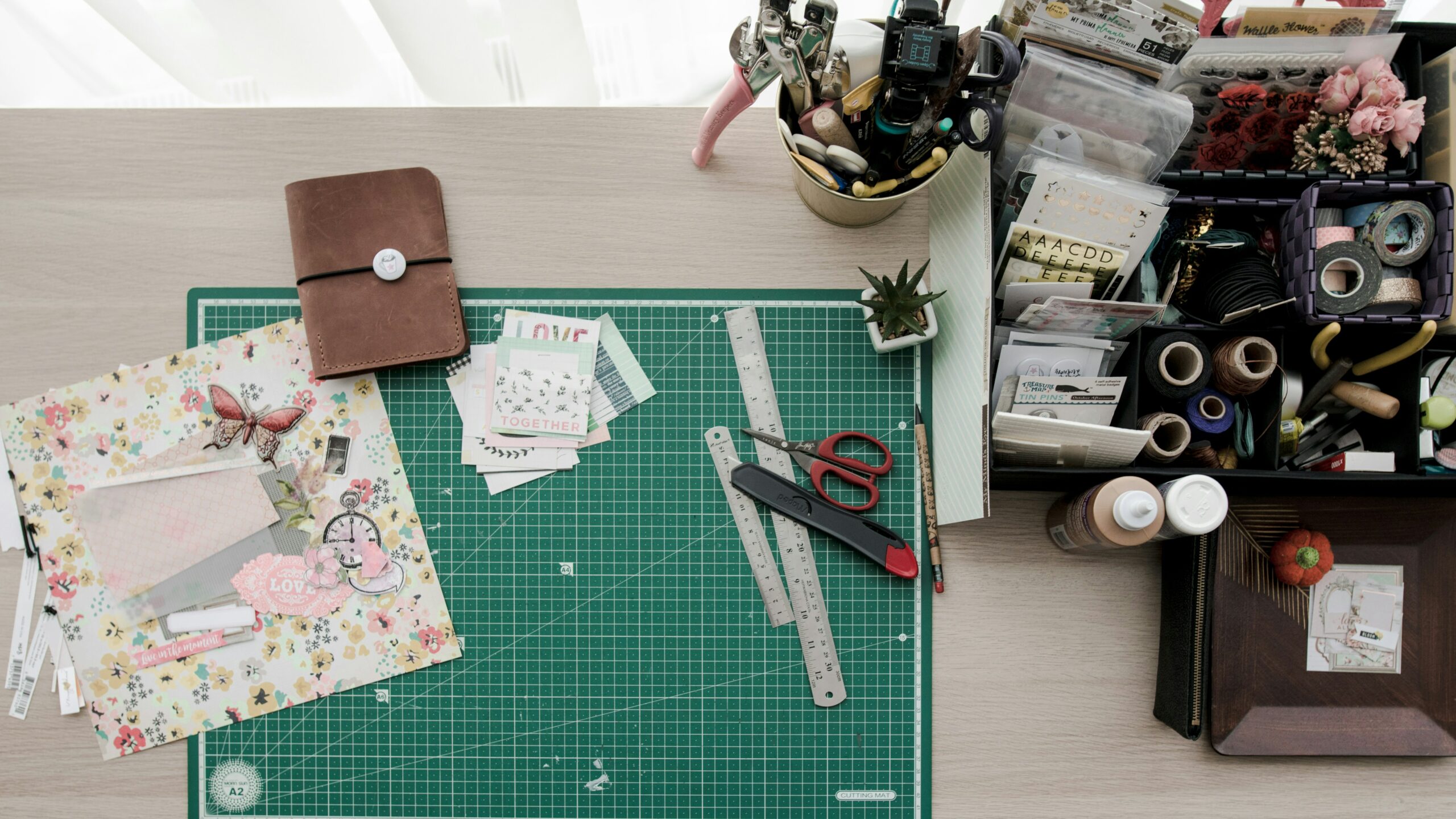 A scrapbook crafting station featuring cutting mats, rulers, scissors, decorative paper, stickers, and various organizational containers filled with crafting supplies.