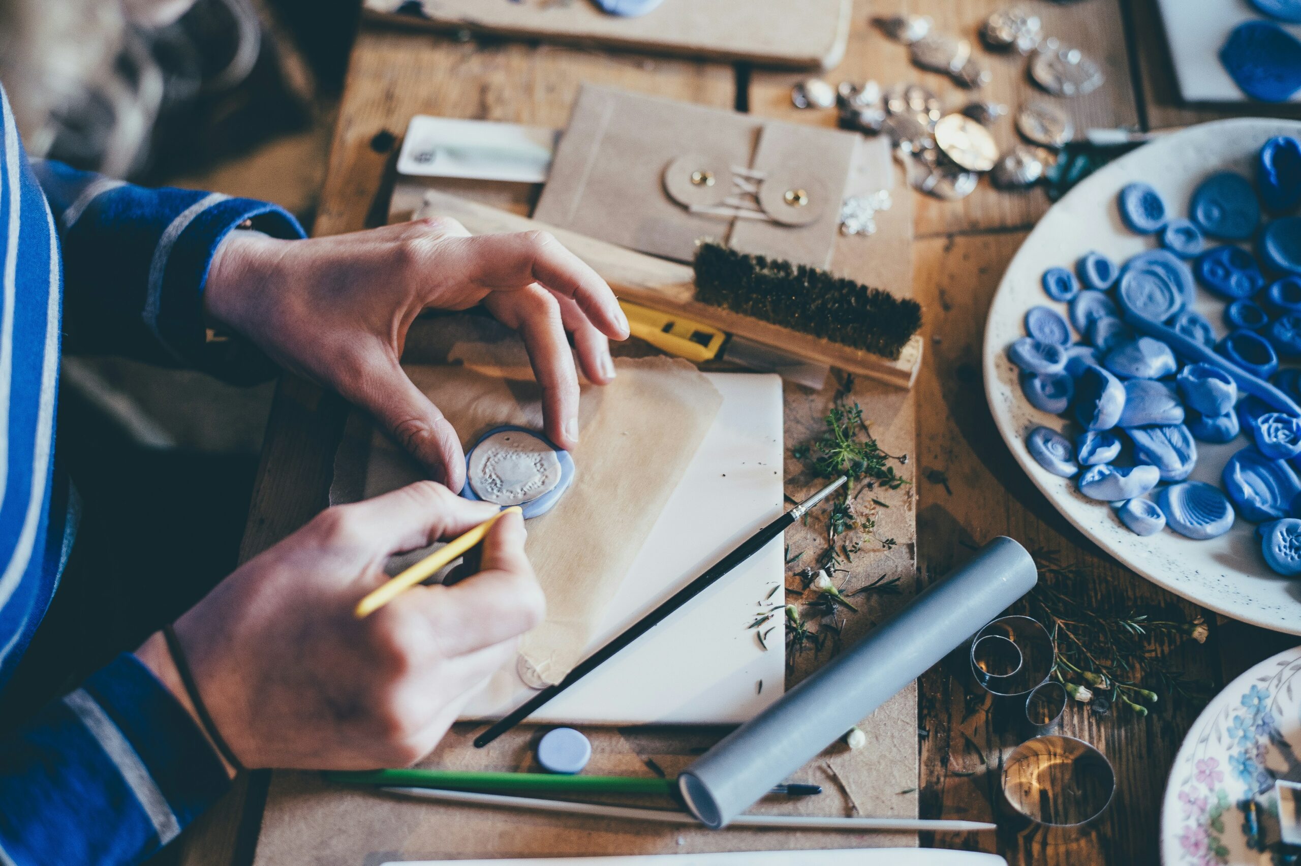 A person working on crafting wax seals, surrounded by various tools and materials such as brushes, wax molds, and decorative items on a wooden table.