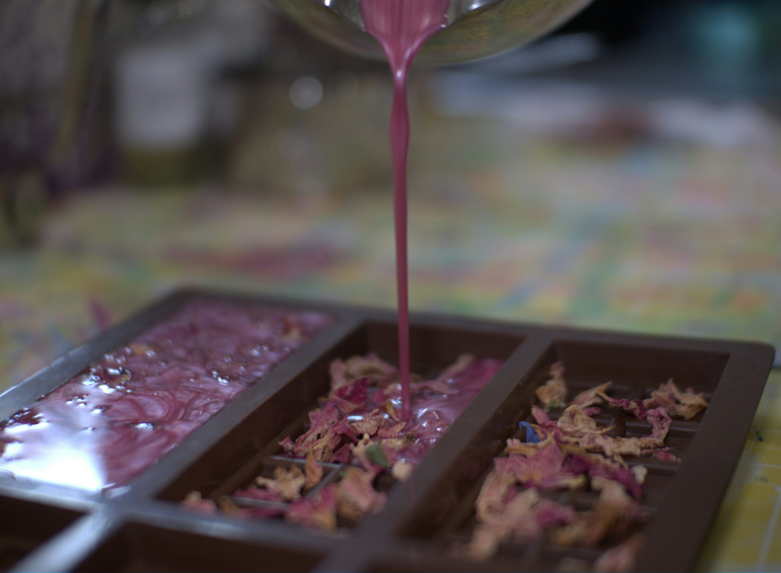 Pink melted wax being poured into a rectangular mold containing dried flower petals, creating handmade wax melts.