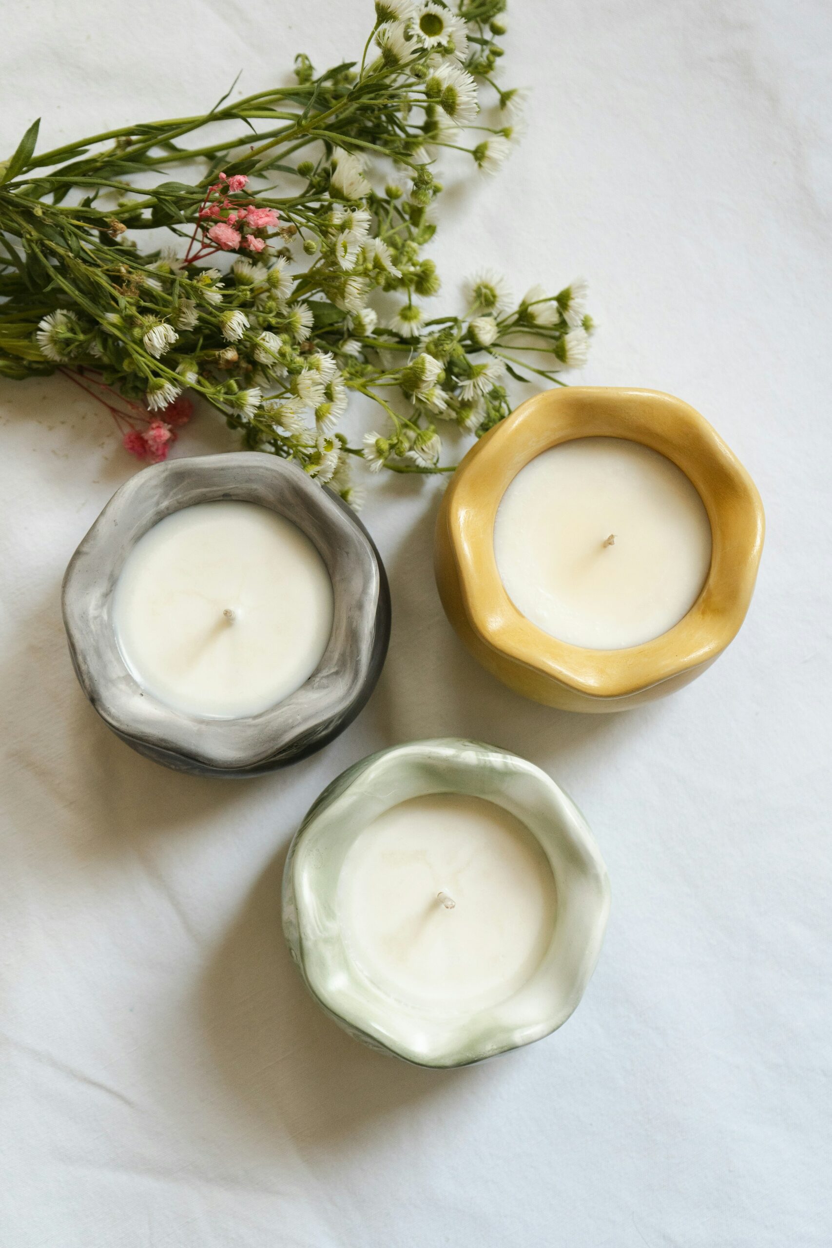 Three handmade candles in uniquely shaped ceramic holders (one yellow, one gray, one light green) are arranged on a white surface next to a small bouquet of white and pink wildflowers.