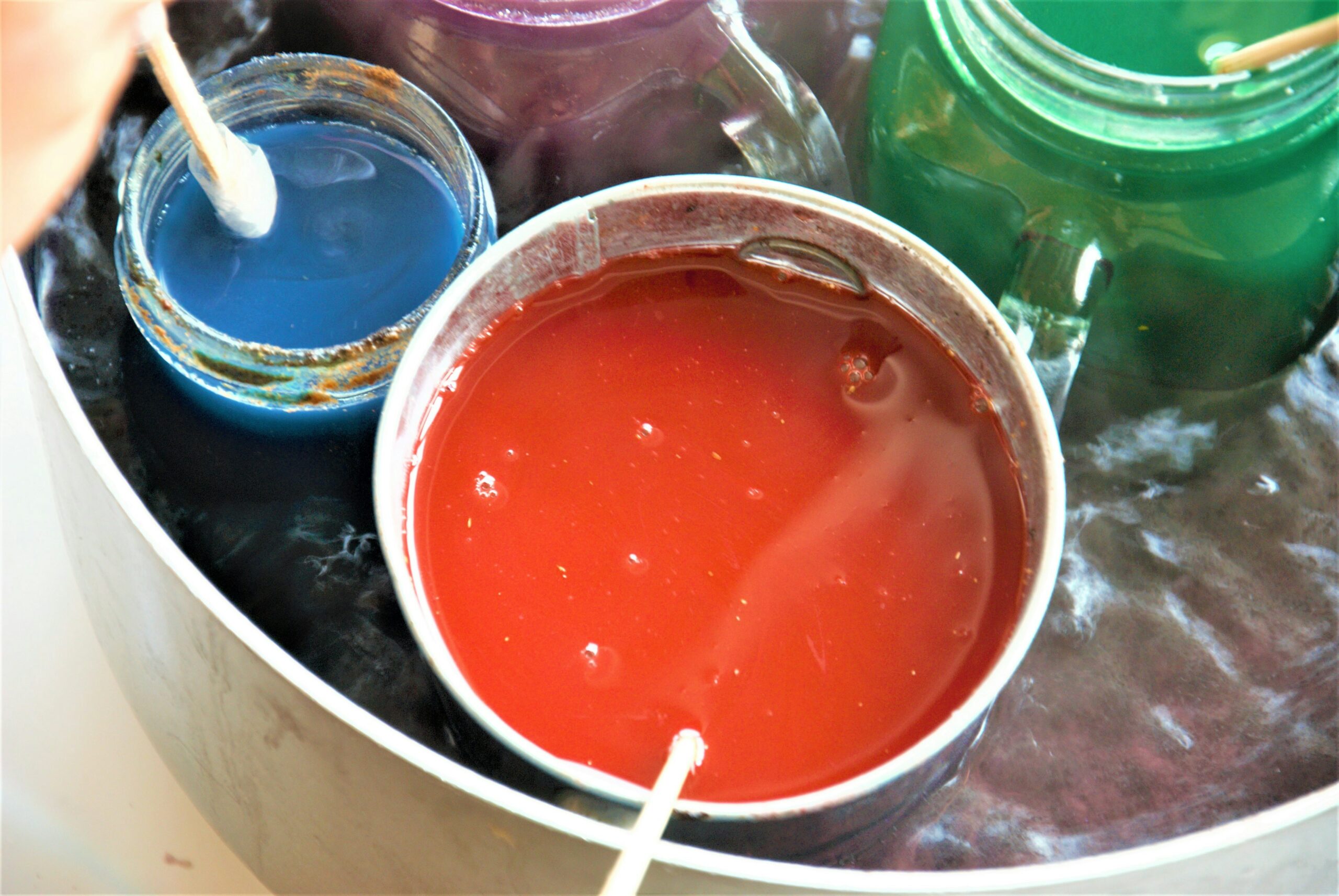 Various jars and containers of colored wax (blue, red, green, purple) melting in a hot water bath on a stove, with wooden sticks for stirring.
