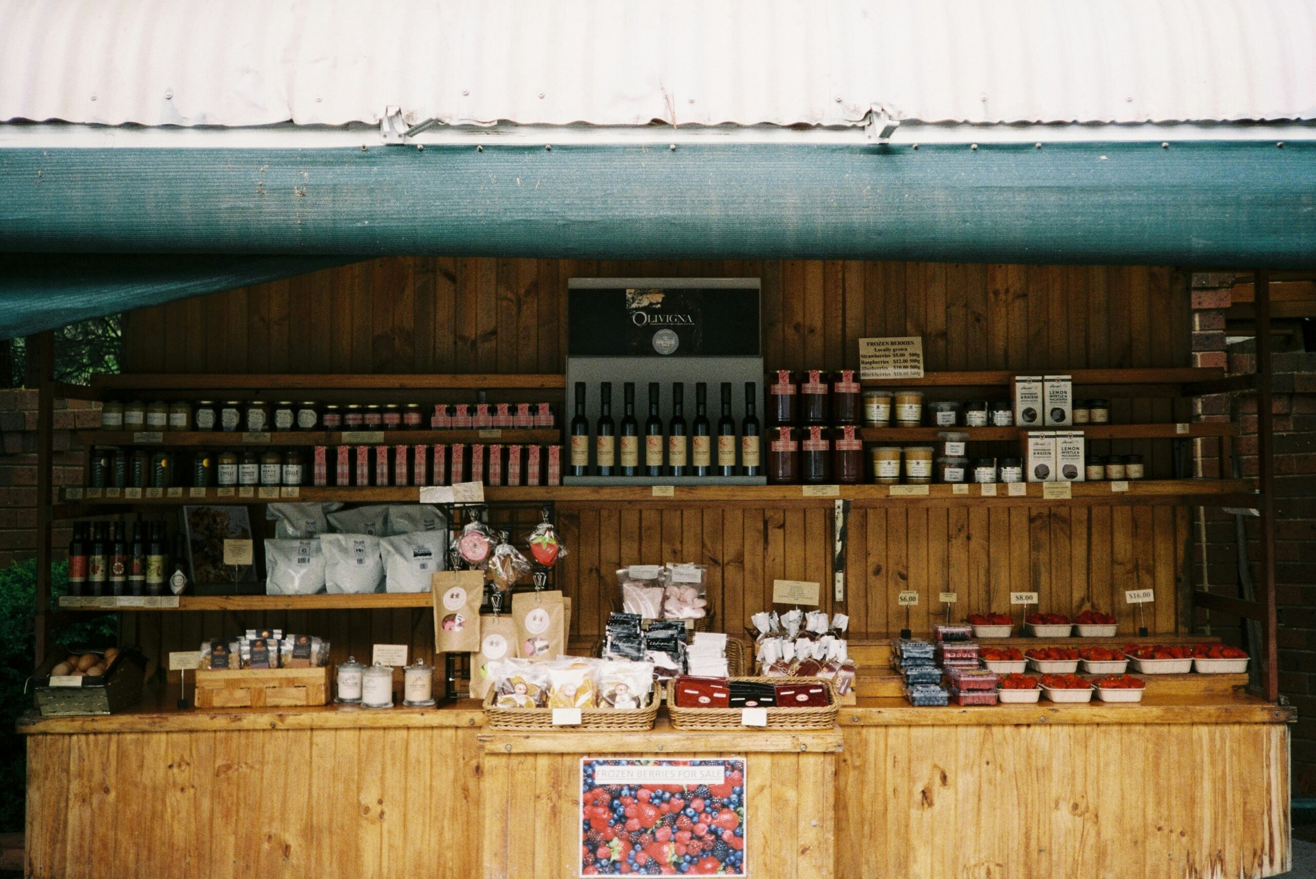 Wooden market stall selling an assortment of packaged goods, bottles, jars, and fresh berries displayed neatly on shelves and counters. The stall is shaded with a green tarp, and a sign on the front advertises frozen berries for sale.