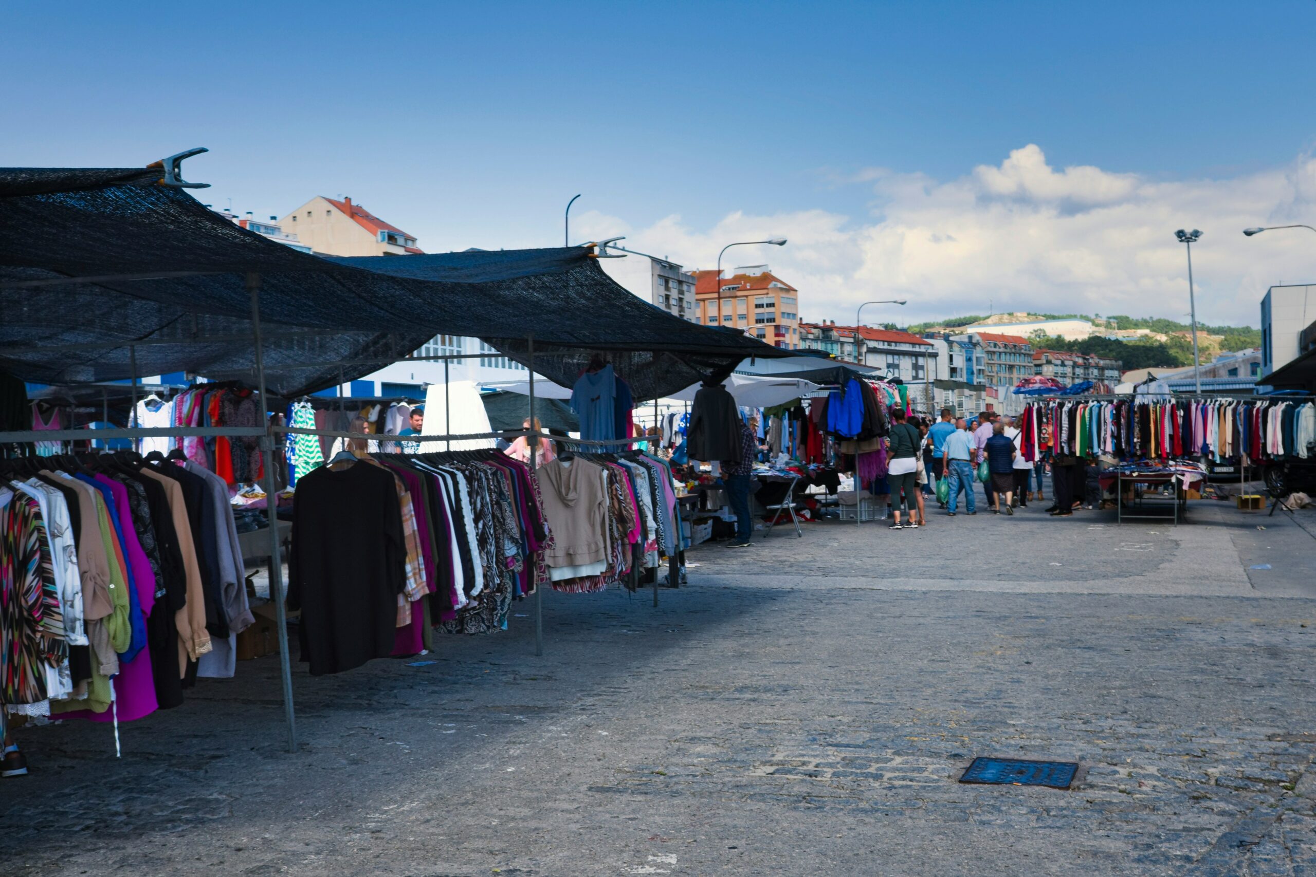Open-air clothing market with rows of racks displaying various colorful garments under black mesh canopies. Shoppers walk and browse the stalls on a sunny day with buildings and hills visible in the distance.