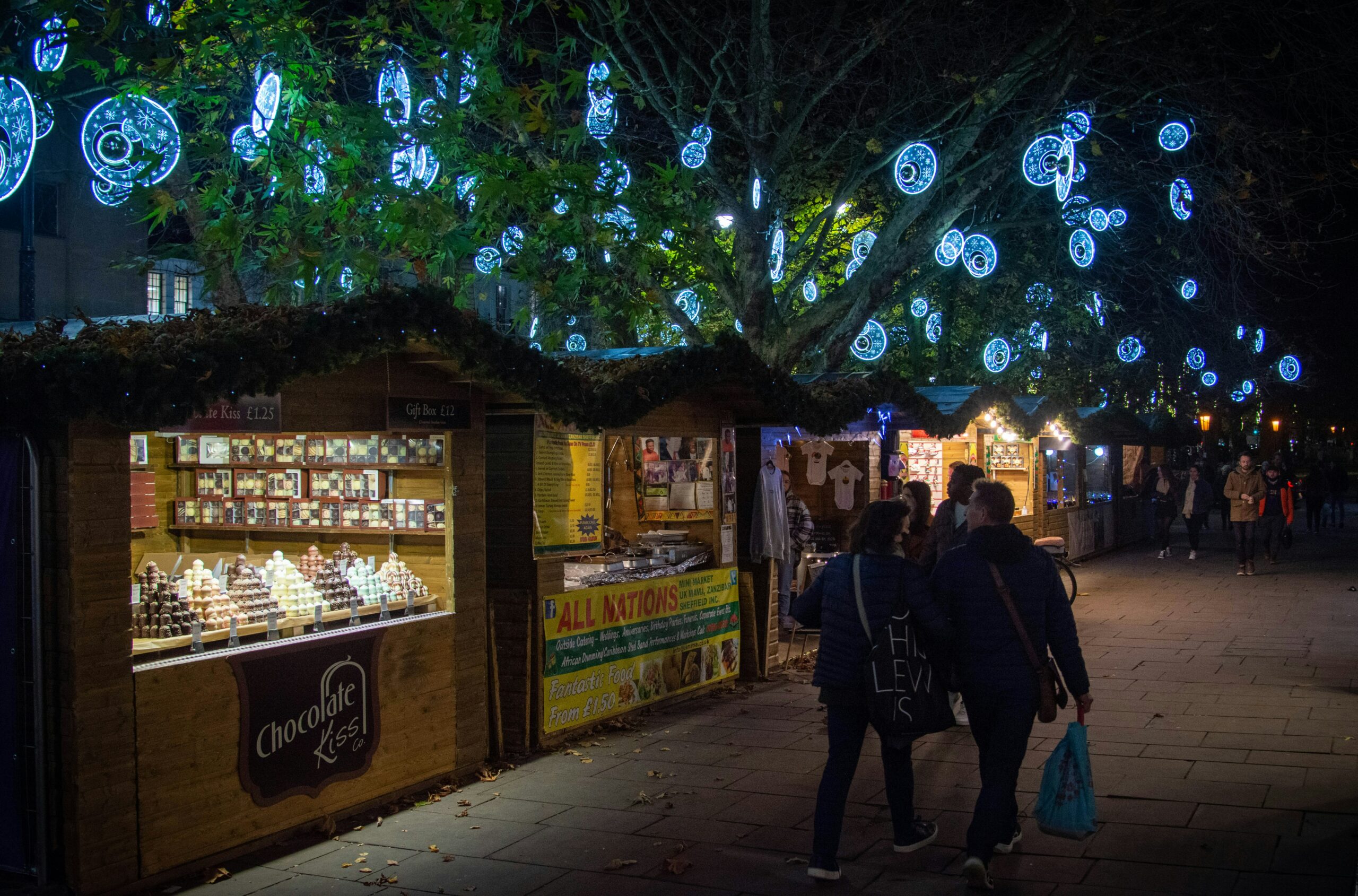 A festive outdoor market at night, with illuminated stalls selling chocolates and other goods, decorated with glowing blue and white circular lights hanging from tree branches. People are walking along the market path.