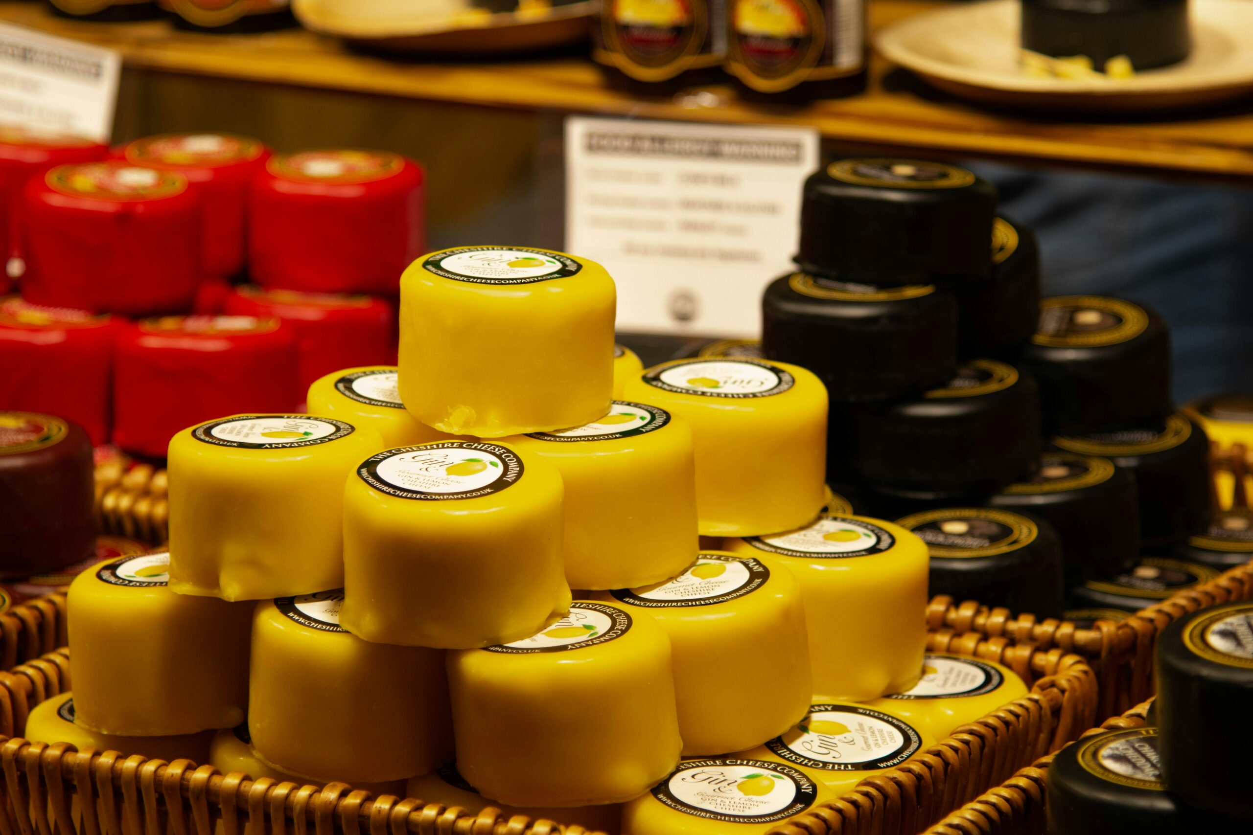 Close-up of small, round cheeses coated in red, yellow, and black wax, stacked in baskets at a market stall. Each cheese has a label on top.