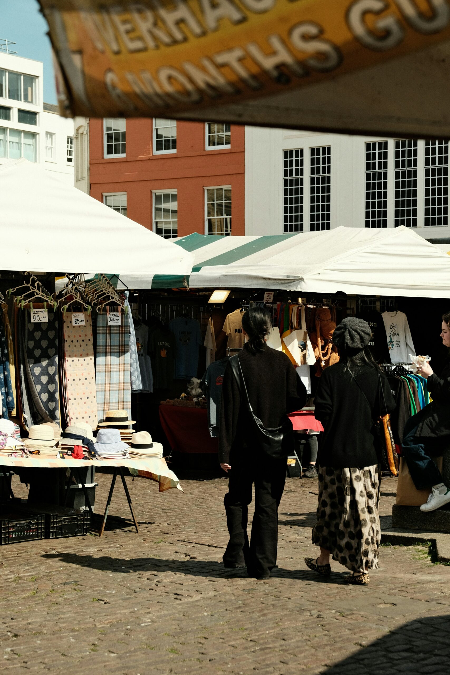 Daytime scene at an outdoor market with white and green striped tents. Clothing, hats, and scarves are displayed for sale, and several people are browsing the stalls on a cobblestone street.