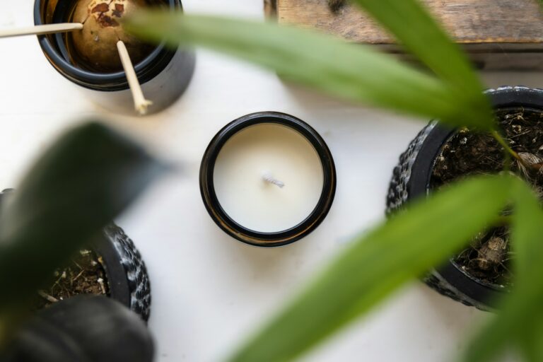 A scented candle, possibly for aromatherapy, with a smooth, white surface. It is lit, and a gentle flame rises from the wick. The background is blurred, suggesting a soft, calming environment.