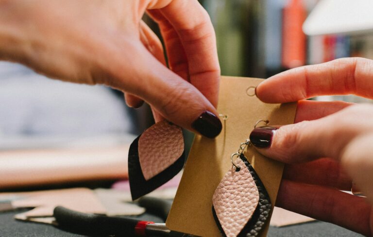 A close-up shot of a person's hands holding a pair of pink and black teardrop-shaped leather earrings, hanging them onto a plain brown cardboard jewellery card.