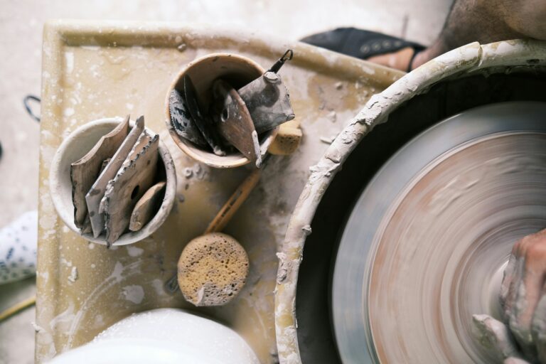 A high-angle close-up shot of a potter's wheel and various pottery tools. The wheel is in motion, blurred slightly by its rotation, and a hand is visible at the bottom right, shaping a piece of clay. To the left of the wheel, a beige bowl holds several flat, irregularly shaped wooden tools, while another cup contains more tools and a sponge. The entire workspace is covered in a fine layer of clay dust and splatters. The scene captures the hands-on, creative process of pottery making.