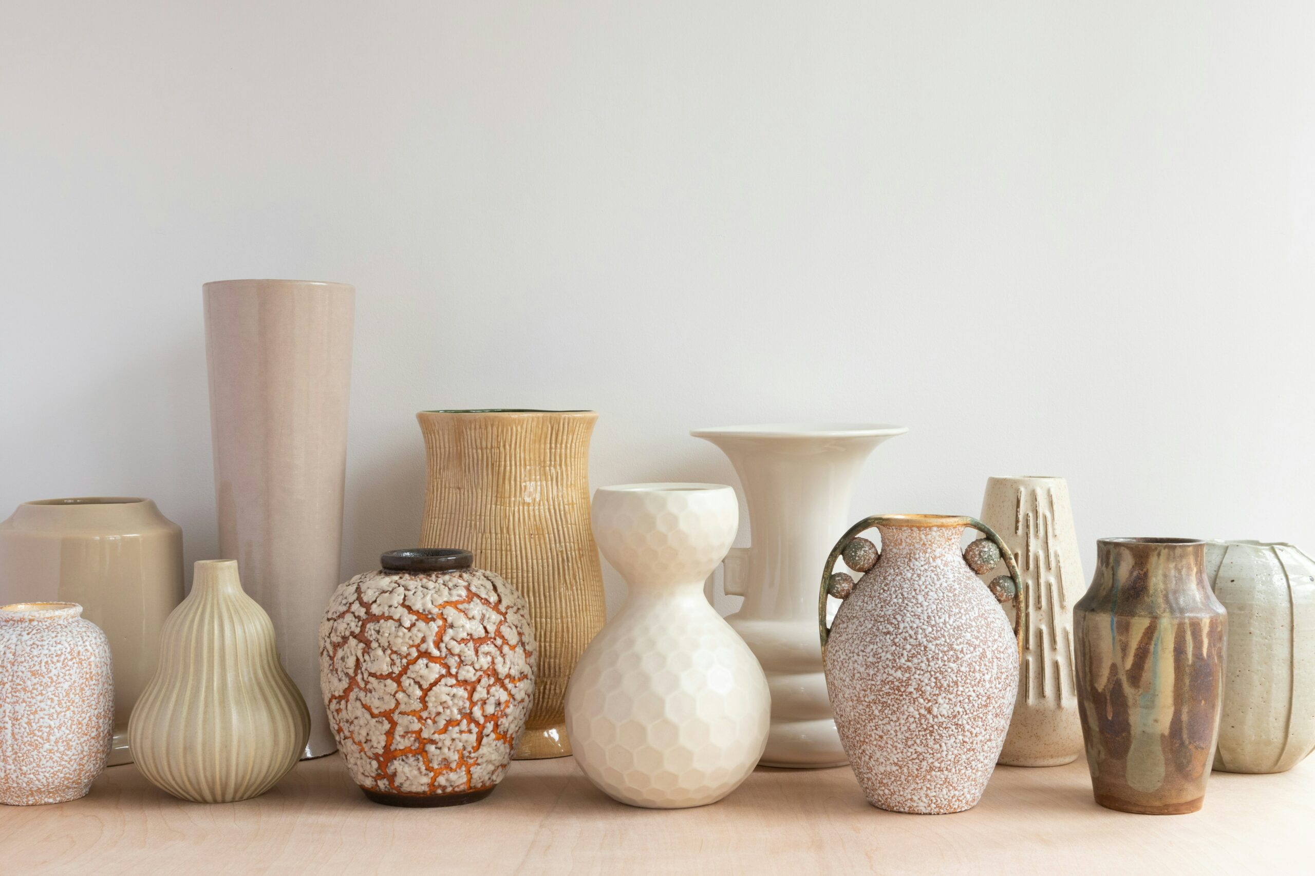 A collection of ceramic vases of various shapes, sizes, and textures sit on a wooden surface against a plain white wall. The vases are arranged in a row and display a range of natural and earthy tones, including beige, brown, off-white, and muted orange. The textures of the vases vary significantly, from smooth and polished surfaces to ridged, honeycomb, and speckled finishes.