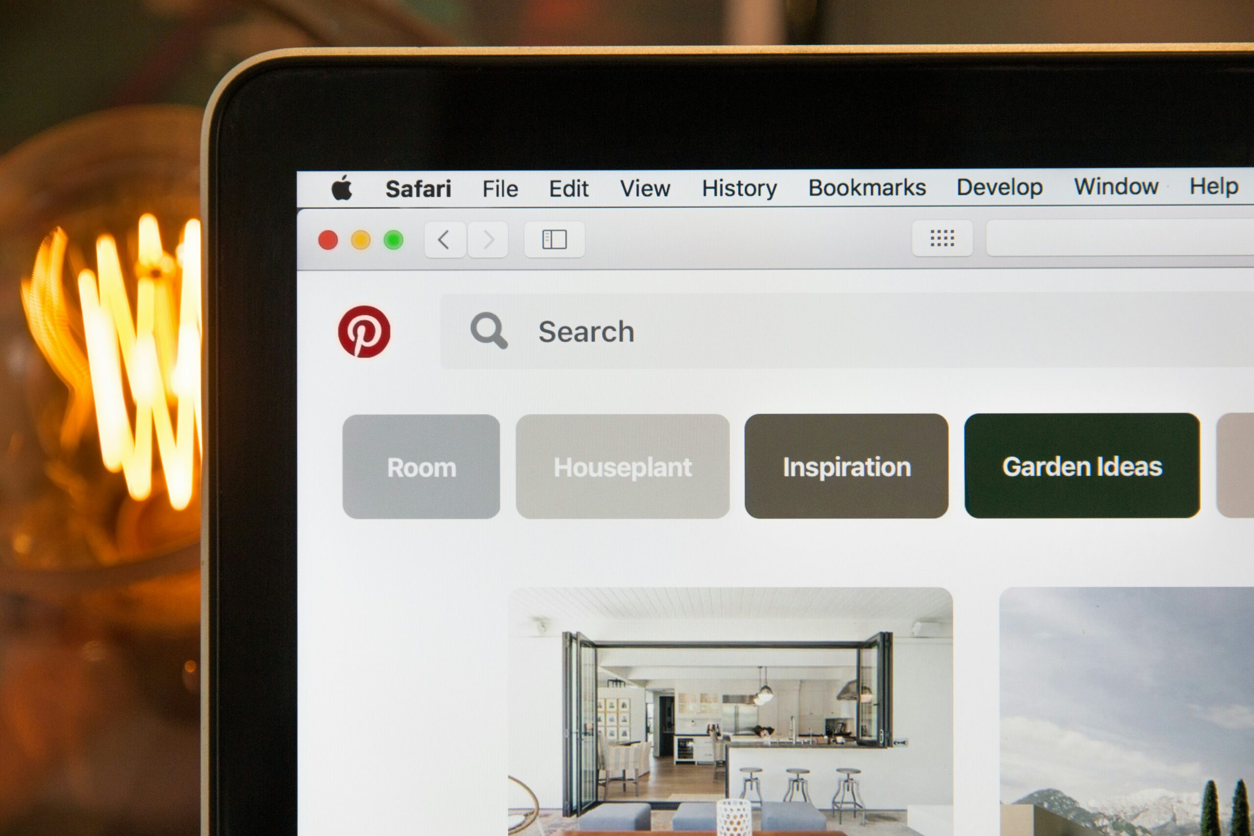  A close-up, eye-level shot shows a person on a laptop looking at the Pinterest website. The screen displays the Safari browser with a search bar and various suggestion buttons like "Room," "Houseplant," "Inspiration," and "Garden Ideas." A section of an image showing a modern kitchen is visible below the buttons. The screen's surface reflects an orange glowing light bulb on the left, which is slightly out of focus.