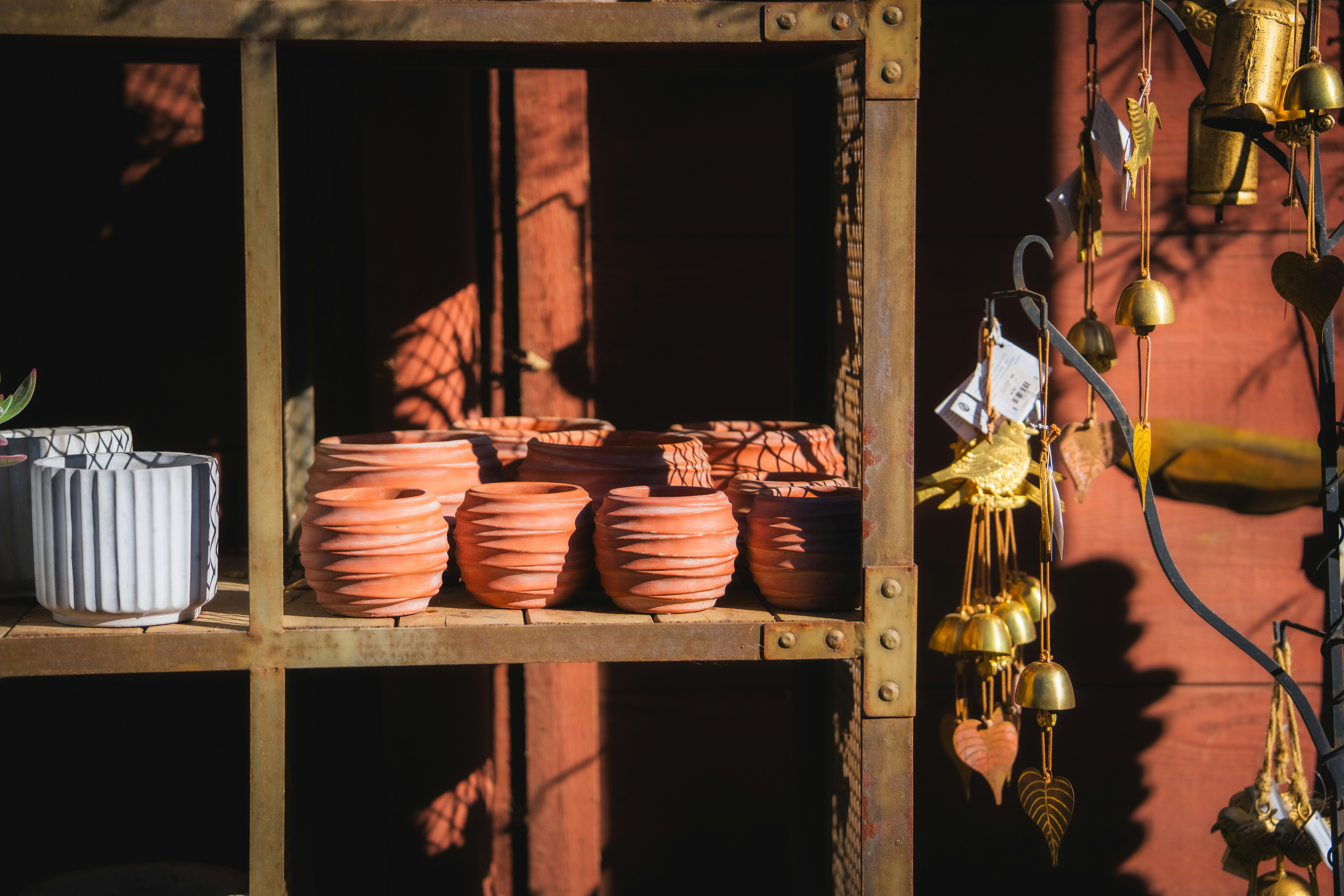  A close-up, eye-level shot shows rows of small, unglazed terracotta pots on a dark brown metal shelf. To the left, a single ridged, white ceramic pot stands out. The background features a reddish-brown wall with diagonal wood paneling. The pots and shelf are bathed in sunlight, while the background and the lower shelves are in shadow. To the right, hanging from a thin black rod, are strings of small golden bells and stylized leaf and bird ornaments.